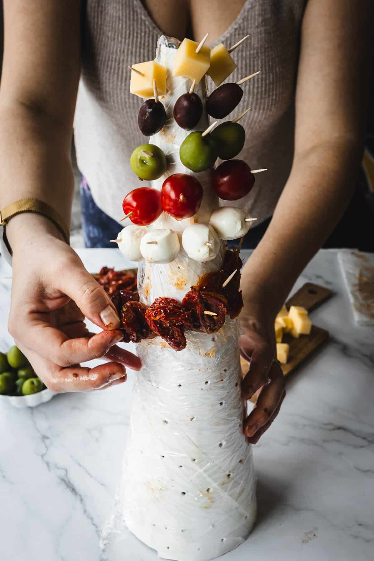 A person arranges skewered ingredients on a conical structure. The skewers hold cheese cubes, olives, cherry tomatoes, and mozzarella balls, with sun-dried tomatoes at the bottom. The setup is on a marble countertop.