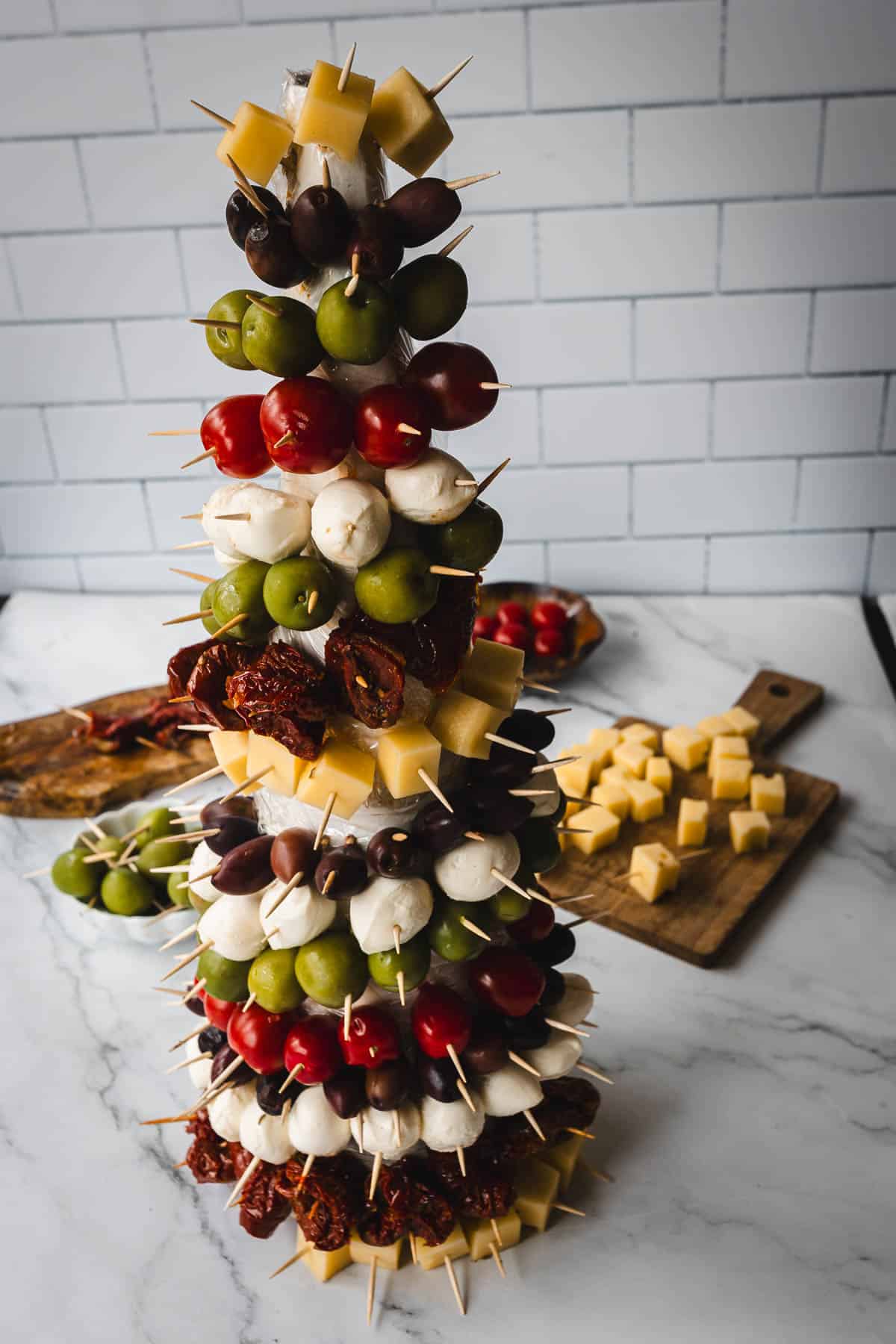 A vertical skewer tower with layers of cheese cubes, black and green olives, cherry tomatoes, and mozzarella balls. The skewers are arranged in a stack on a marble surface with additional cheese cubes on a wooden board in the background.