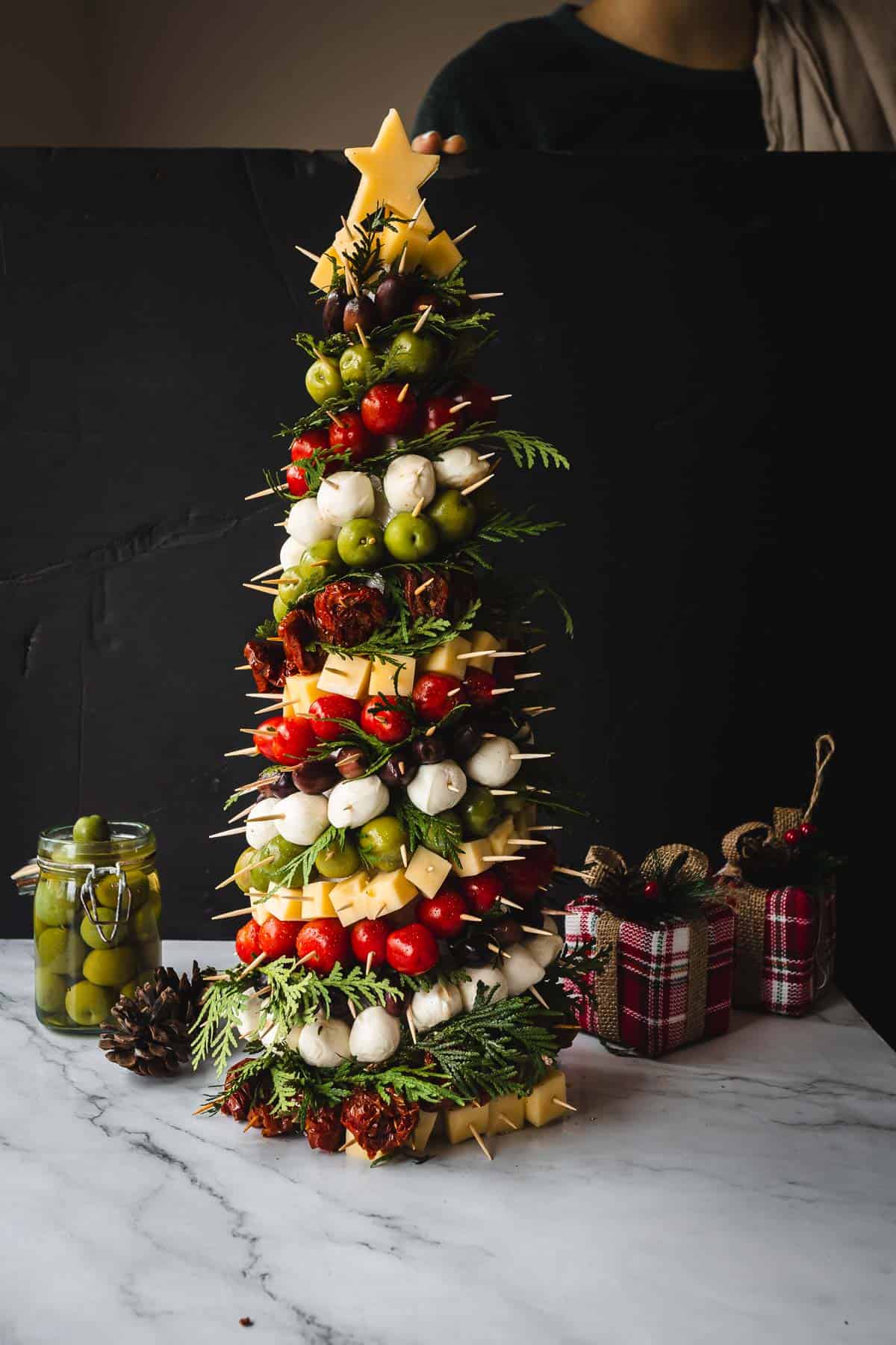 A festive Christmas tree-shaped food arrangement made of skewered cheese cubes, tomatoes, olives, and greens sits on a marble countertop. A jar of olives is on the left, and wrapped gifts are on the right. A cheese star tops the tree.