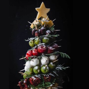 A festive tree-shaped arrangement made of assorted vegetables and cheeses is topped with a star-shaped cheese. It includes olives, cherry tomatoes, bocconcini, and is decorated with sprigs on a dark background.