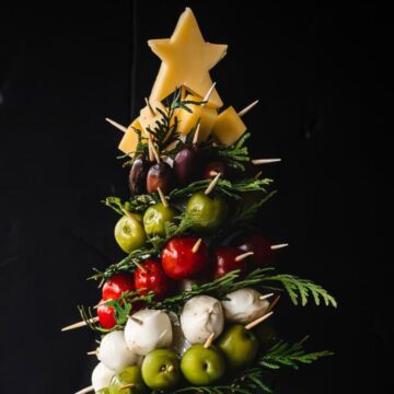 A food arrangement resembling a Christmas tree features layers of olives, cherry tomatoes, mozzarella balls, and herbs, topped with cheese in the shape of a star. Skewers hold the ingredients together against a dark background.