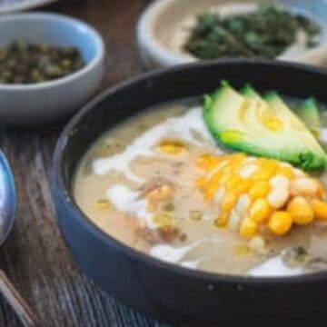 A bowl of soup sits on a wooden table, reminiscent of Grandma's secret recipes. It's garnished with a slice of corn on the cob, avocado slices, and a drizzle of cream. Two spoons rest beside it, with small dishes containing herbs and capers in the background.