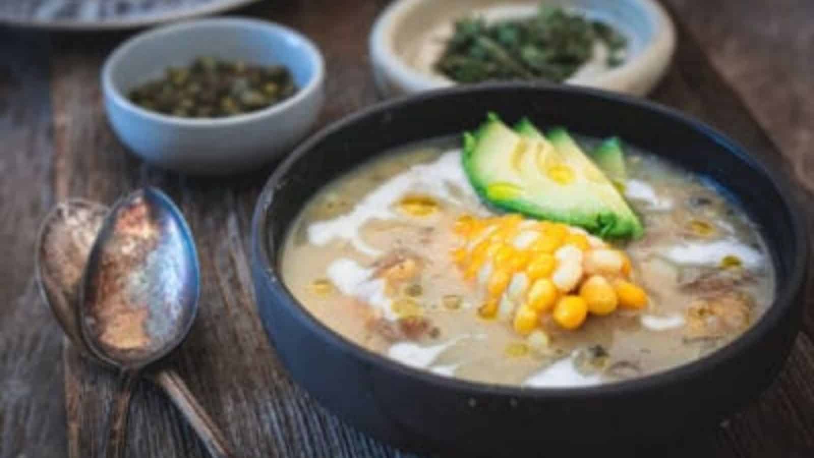 A bowl of soup sits on a wooden table, reminiscent of Grandma's secret recipes. It's garnished with a slice of corn on the cob, avocado slices, and a drizzle of cream. Two spoons rest beside it, with small dishes containing herbs and capers in the background.