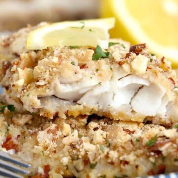 A plate with breadcrumb-crusted fish garnished with lemon slices and herbs. A fork rests beside the fish. A halved lemon is visible in the background.