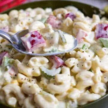 A bowl of macaroni salad containing elbow pasta, diced red bell peppers, celery, and a creamy dressing. A spoon is lifting a portion from the bowl. Sliced pickles are visible in the background.