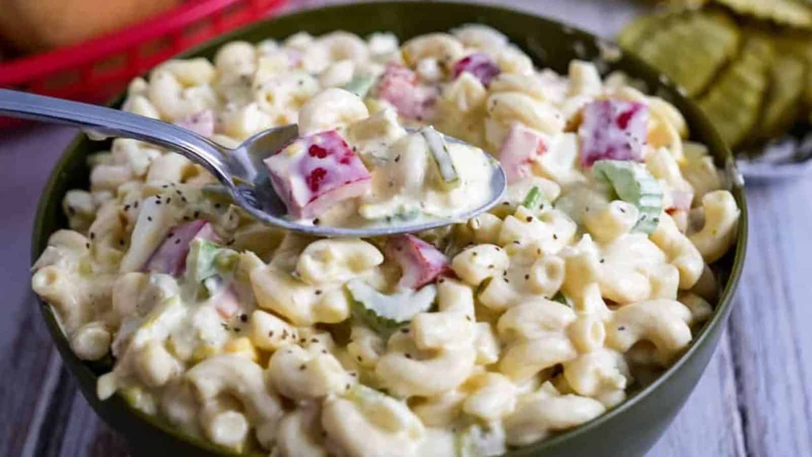 A bowl of macaroni salad containing elbow pasta, diced red bell peppers, celery, and a creamy dressing. A spoon is lifting a portion from the bowl. Sliced pickles are visible in the background.