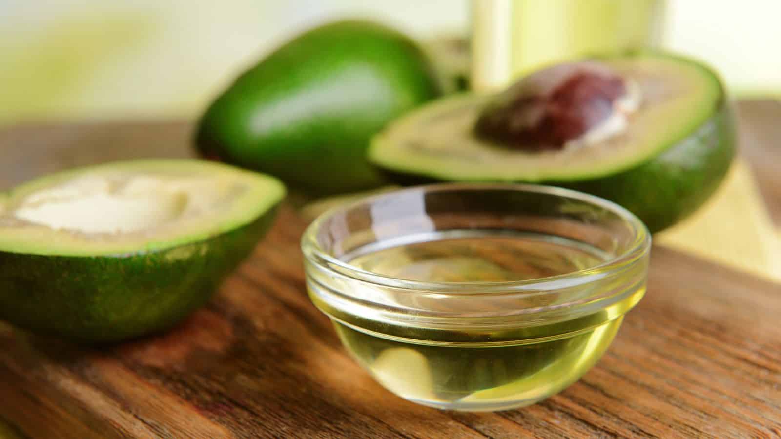 A small glass bowl of healthy oils sits on a wooden cutting board. Behind it, two avocados are visible, one cut in half with the pit exposed. The background is softly blurred, featuring a light green and yellow hue, adding a serene touch.