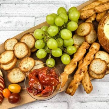 A wooden platter with sliced bread, grape tomatoes, sliced apples, green grapes, roasted tomatoes, breadsticks, and a round loaf of bread filled with melted cheese, garnished with herbs. The background is a white brick surface.
