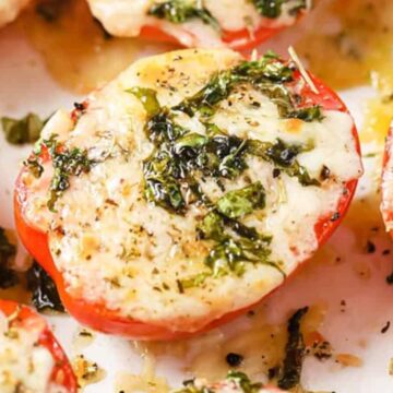 Close-up of sliced tomatoes topped with melted cheese, herbs, and seasoning. The tomatoes are arranged on a baking sheet, showcasing a golden, bubbling surface with bits of green herbs and a sprinkle of pepper.