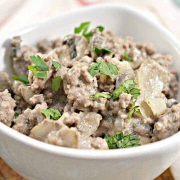 A white bowl filled with a ground meat mixture, likely beef, garnished with chopped parsley evokes the charm of old-school dishes. The meal includes onion and possibly mushrooms. A white cloth with a red stripe adds to the nostalgic feel in the background.