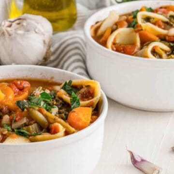 Two bowls of vegetable soup with pasta, garnished with parsley, sit on a white surface. A head of garlic, garlic cloves, olive oil, and a striped cloth are in the background. Spoons are placed in each bowl, reminiscent of comforting recipes from the '60s enjoyed by Baby Boomers.
