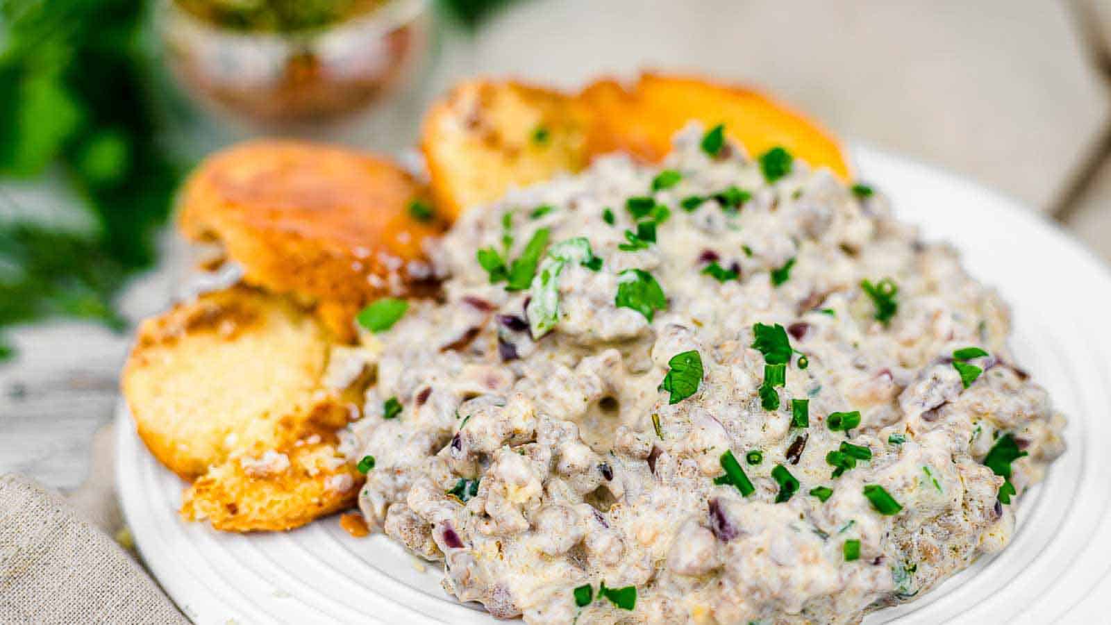 A white plate with a creamy sausage and gravy mixture topped with chopped herbs. On the side are slices of toasted garlic bread. The background features a blurred setting with greens.