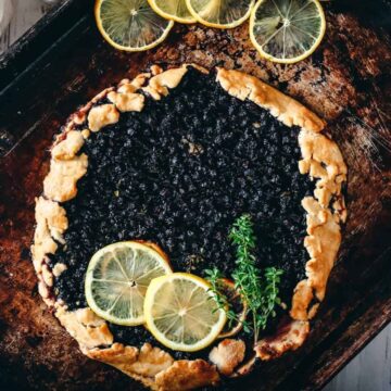 A rustic blueberry galette topped with lemon slices and a sprig of thyme sits on a weathered baking sheet. The crust is golden brown, and the galette is placed on a wooden surface with a light-colored cloth nearby.