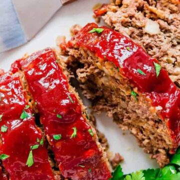 A sliced meatloaf topped with a glossy ketchup glaze is displayed on a white plate. A serving spatula is placed beside it. The meatloaf is garnished with chopped parsley, adding a touch of green.