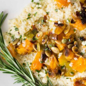 A plate of couscous topped with roasted squash, pumpkin seeds, and herbs. Rosemary sprigs garnish the side. The couscous mixture appears well-seasoned and colorful, served on a white dish placed on a wooden surface.