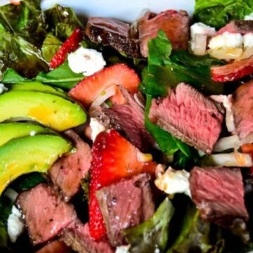 A salad in a bowl featuring sliced avocado, strawberries, pieces of steak, crumbled cheese, and fresh leafy greens.