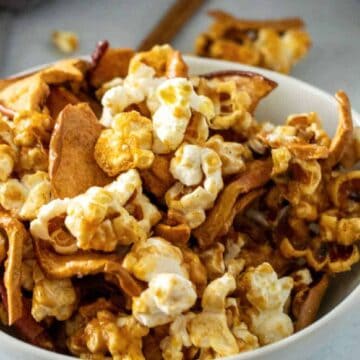 A bowl filled with popcorn mixed with caramel-coated apple slices sits on a light-colored surface, perfect for those happy weekend movie nights. Some of the delicious snacks have spilled from the bowl, and a gray cloth is partly visible on the side.