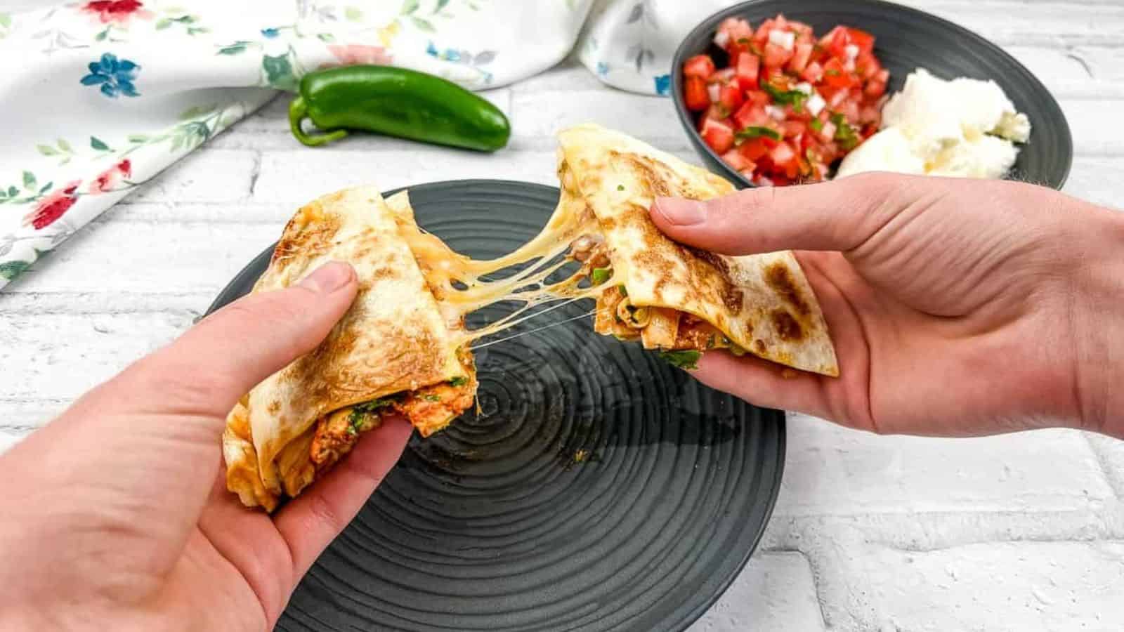 Two hands pull apart a cheesy quesadilla, revealing delightful cheese stretches. A green pepper, white napkin with a floral pattern, and bowls of diced tomatoes and cheese adorn the background on a white surface—a perfect choice for kid-friendly recipes that even the pickiest eaters will love.