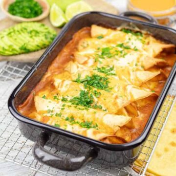A family baking dish of enchiladas covered with melted cheese and garnished with chopped herbs. Slices of avocado, lime wedges, and a small bowl of love-infused herbs are on the side, along with a stack of hearty tortillas on a wire rack. A glass of orange juice completes the scene.