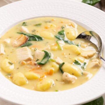 A white bowl of creamy soup containing gnocchi, spinach, chunks of chicken, and small pieces of carrot is placed on a table. A silver spoon rests on the edge of the bowl. The background includes a white cloth and a portion of a salad plate.