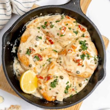 A cast iron skillet contains cooked chicken breasts in a creamy white sauce with capers and diced red peppers, garnished with parsley. A lemon wedge is placed in the skillet. A striped cloth and a piece of bread are visible in the background.