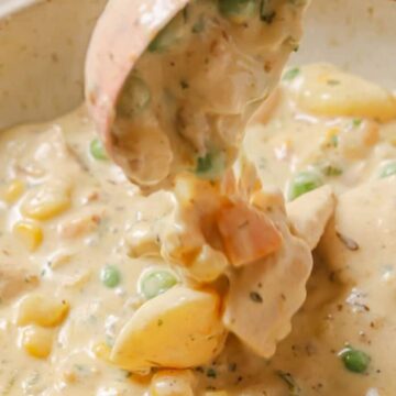 A close-up of a creamy dish containing chunks of chicken, potatoes, peas, and corn. The ingredients are mixed in a thick, beige sauce, and a spoon is lifting a portion from a bowl.