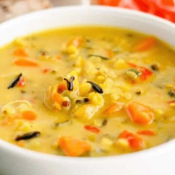 A white bowl filled with creamy yellow soup containing diced vegetables and herbs. The soup appears to have carrots and possibly wild rice or similar grains. In the background, there is a slice of bread and a red pattern cloth.