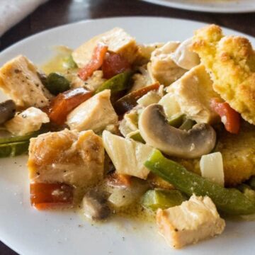 A white plate holds a vibrant meal featuring chicken pieces, green bell peppers, mushrooms, tomatoes, and cornmeal dumplings. The light sauce enhances this old-school dish's timeless appeal. It sits on a wooden table with a fork in the background.