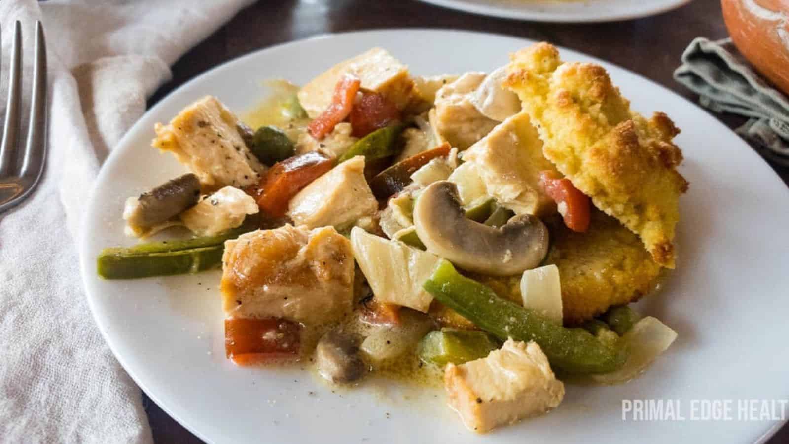 A white plate holds a vibrant meal featuring chicken pieces, green bell peppers, mushrooms, tomatoes, and cornmeal dumplings. The light sauce enhances this old-school dish's timeless appeal. It sits on a wooden table with a fork in the background.