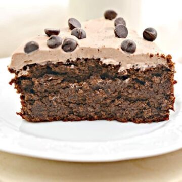A slice of chocolate cake with a layer of frosting, topped with chocolate chips, is placed on a white plate. The cake appears moist and rich, sitting on a light wooden surface.