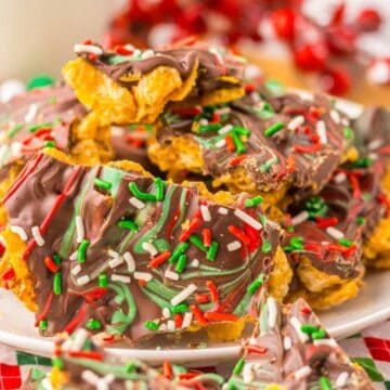 A plate of chocolate bark is topped with red, green, and white sprinkles. The treats are arranged on a white plate with a colorful, festive backdrop.