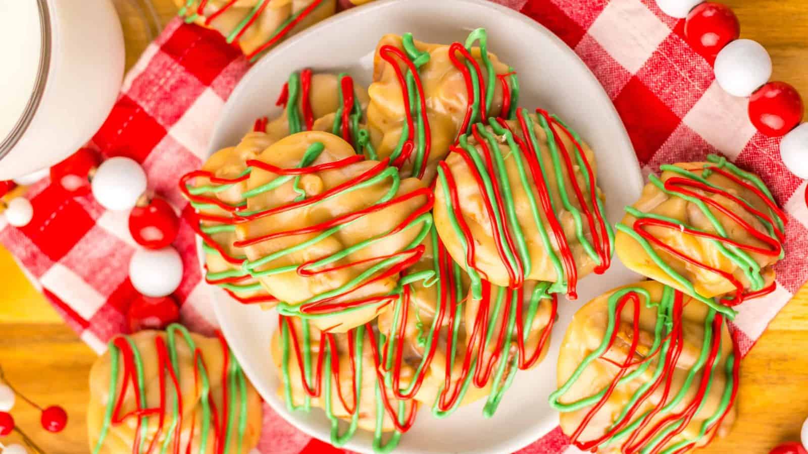 A plate of white chocolate-covered treats drizzled with red and green icing sits on a red and white checkered cloth. A glass of milk is visible nearby, and colorful spherical decorations surround the arrangement.