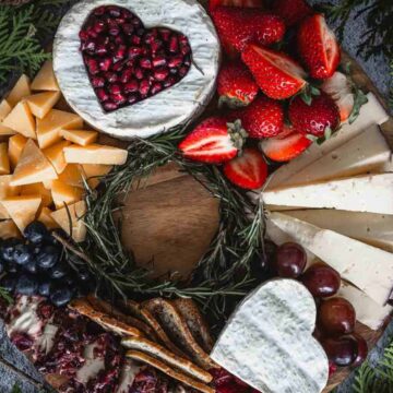 A charcuterie board with heart-shaped cheese topped with pomegranate seeds, surrounded by sliced strawberries, blueberries, grapes, assorted cheeses, crackers, and pine boughs.