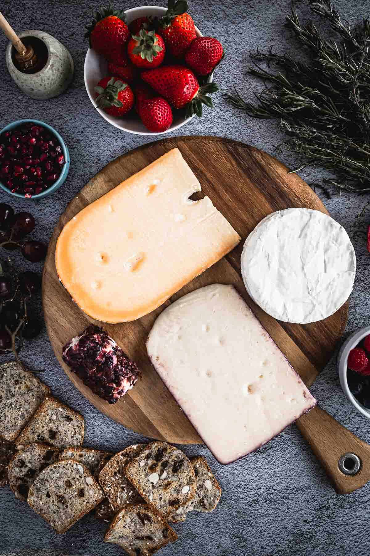 A wooden board displays three types of cheese, including a round brie, a block with a slice cut from it, and a dark-rinded piece. Nearby are bowls of strawberries, berries, sprigs of herbs, and sliced bread.