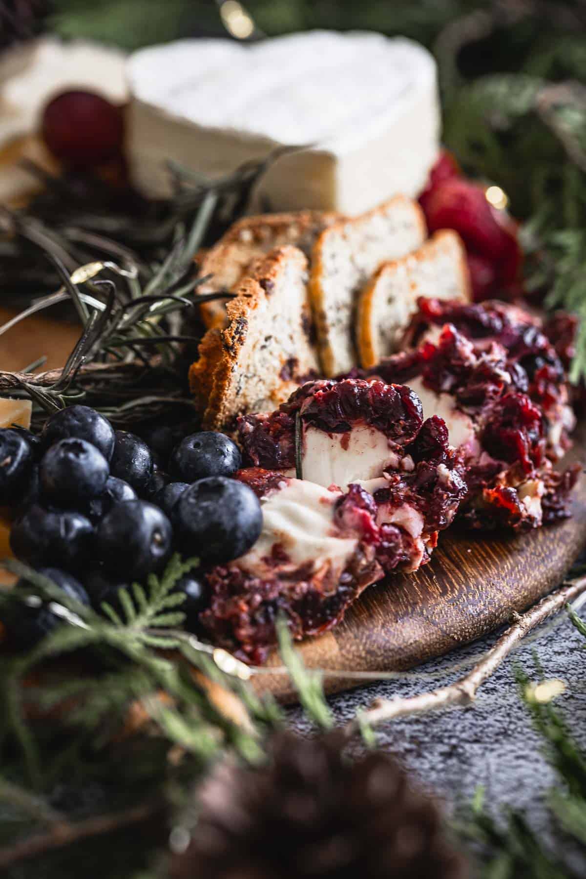 A rustic cheese platter featuring various cheeses, bread slices, a rich red chutney, and a bunch of dark grapes. Decorated with fresh rosemary and seasonal greenery, the platter rests on a wooden surface.