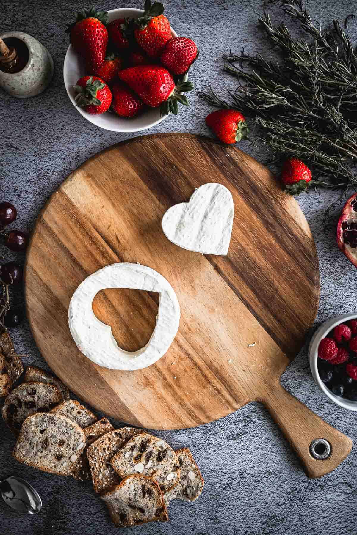 A wooden cutting board holds a circular piece of white cheese with a heart-shaped cut-out and a separate heart-shaped piece of cheese. Surrounding the board are bowls of strawberries, grapes, berries, sprigs of herbs, and slices of bread.