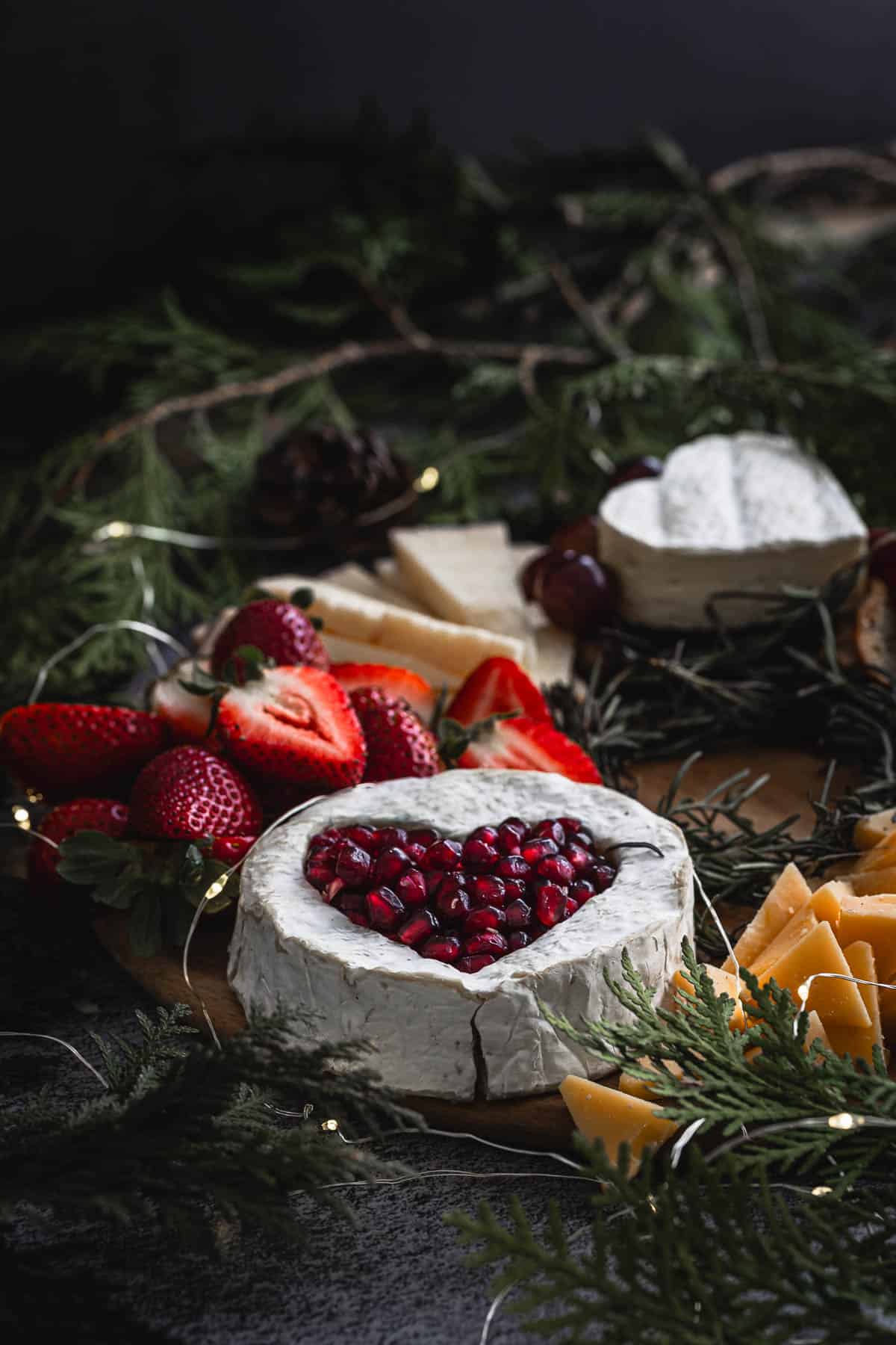 A festive cheese platter featuring a round wheel of brie with a heart-shaped topping of pomegranate seeds. The platter is adorned with slices of cheese, crackers, strawberries, and festive greenery, surrounded by soft decorative lights.