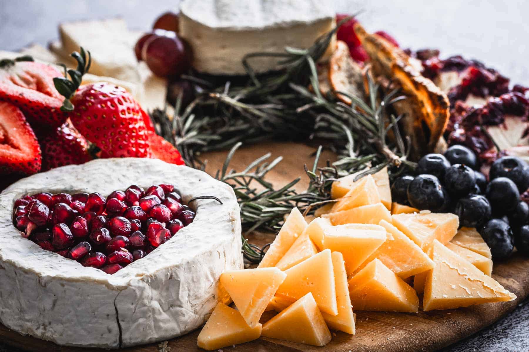 A wooden platter with various cheeses, including a wheel topped with pomegranate seeds, triangular slices of cheese, strawberries, blueberries, and crackers. Rosemary sprigs are scattered for decoration.