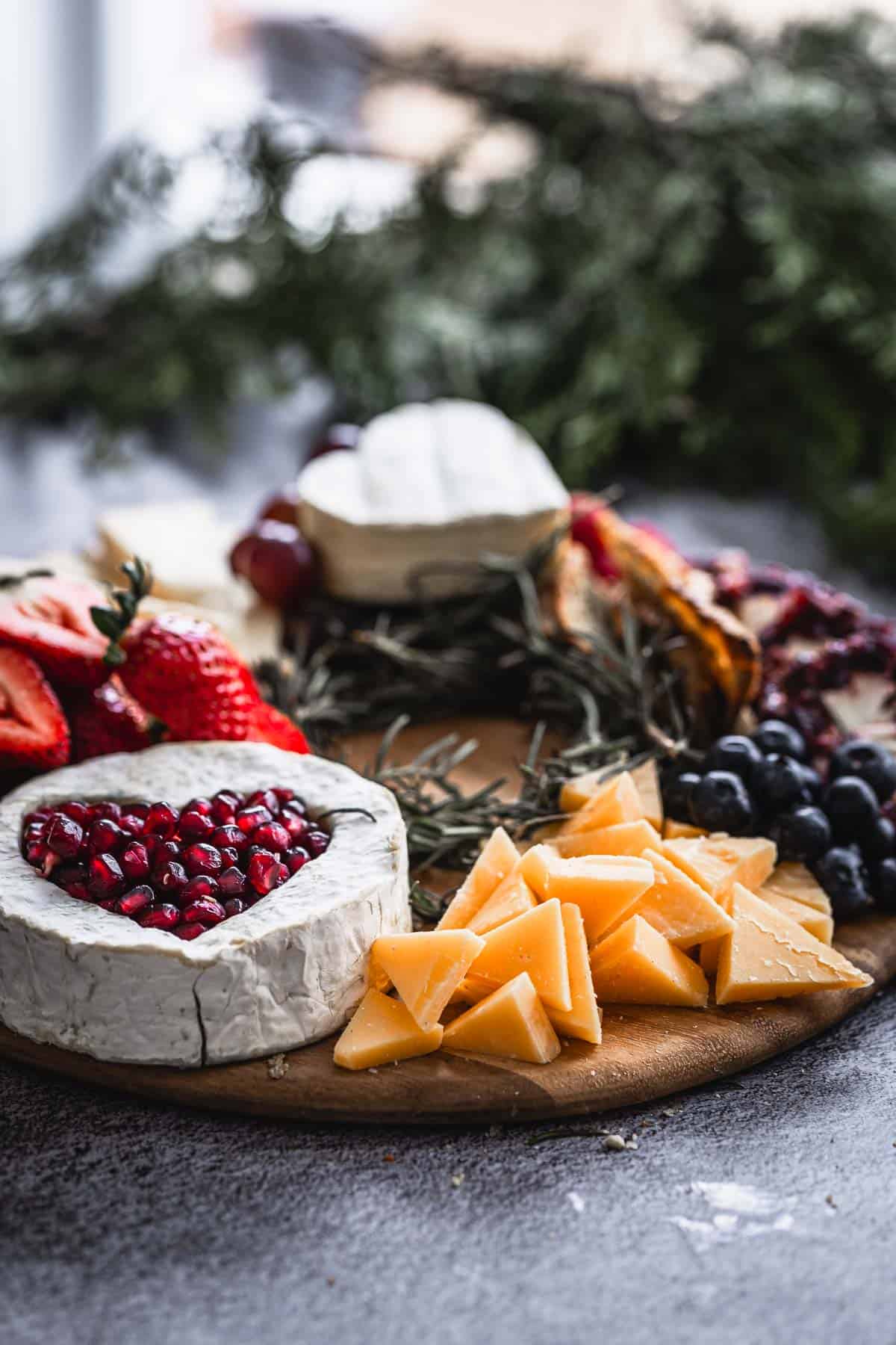 A wooden platter with various cheeses, sliced strawberries, blueberries, pomegranate seeds, and rosemary sprigs. The focus is on a brie cheese topped with pomegranate seeds and sliced cheddar cheese. There are evergreen branches in the background.