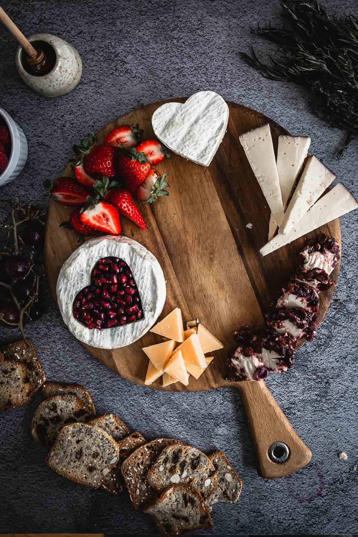 A wooden board displays a variety of foods, including sliced strawberries, heart-shaped brie cheese topped with pomegranate seeds, assorted cheese slices, and a row of round cheese balls with a crust. Nearby are sliced bread and a small vase.