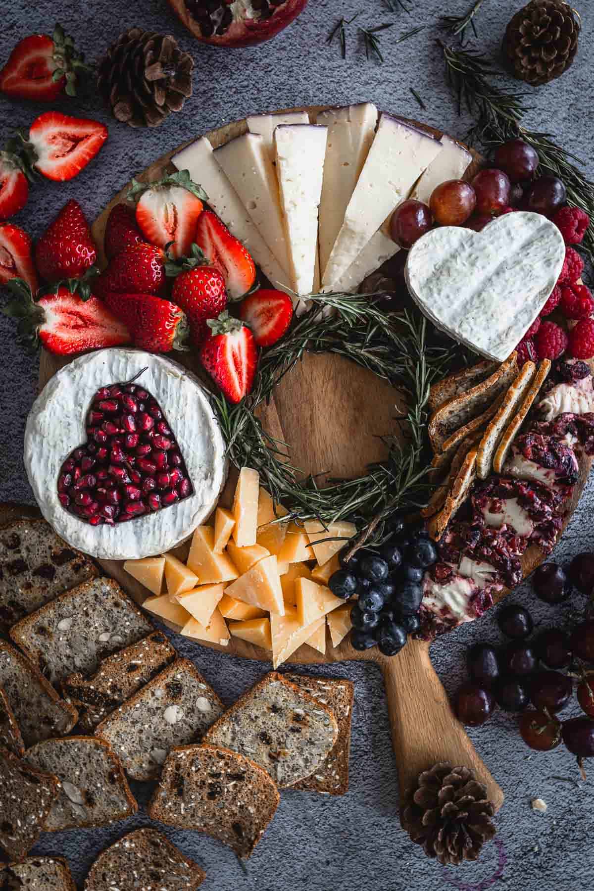 A round wooden board with various cheeses, including a heart-shaped one topped with pomegranate seeds, sliced strawberries, grapes, blueberries, nuts, and whole grain bread slices. Pine cones and rosemary sprigs are used for decoration.