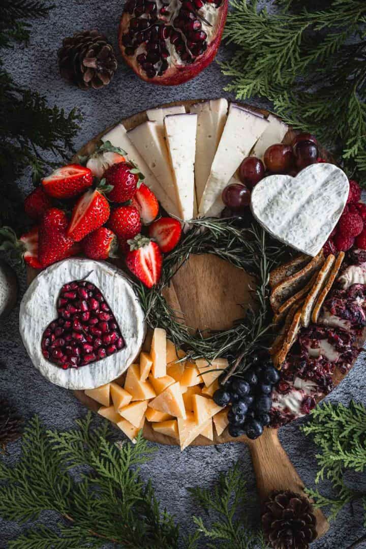 A wooden cheese board with various cheeses, including heart-shaped ones topped with pomegranate seeds, surrounded by strawberries, grapes, sliced bread, and pine branches. Pinecones decorate the edges, and small cheese cubes are gathered near the center.