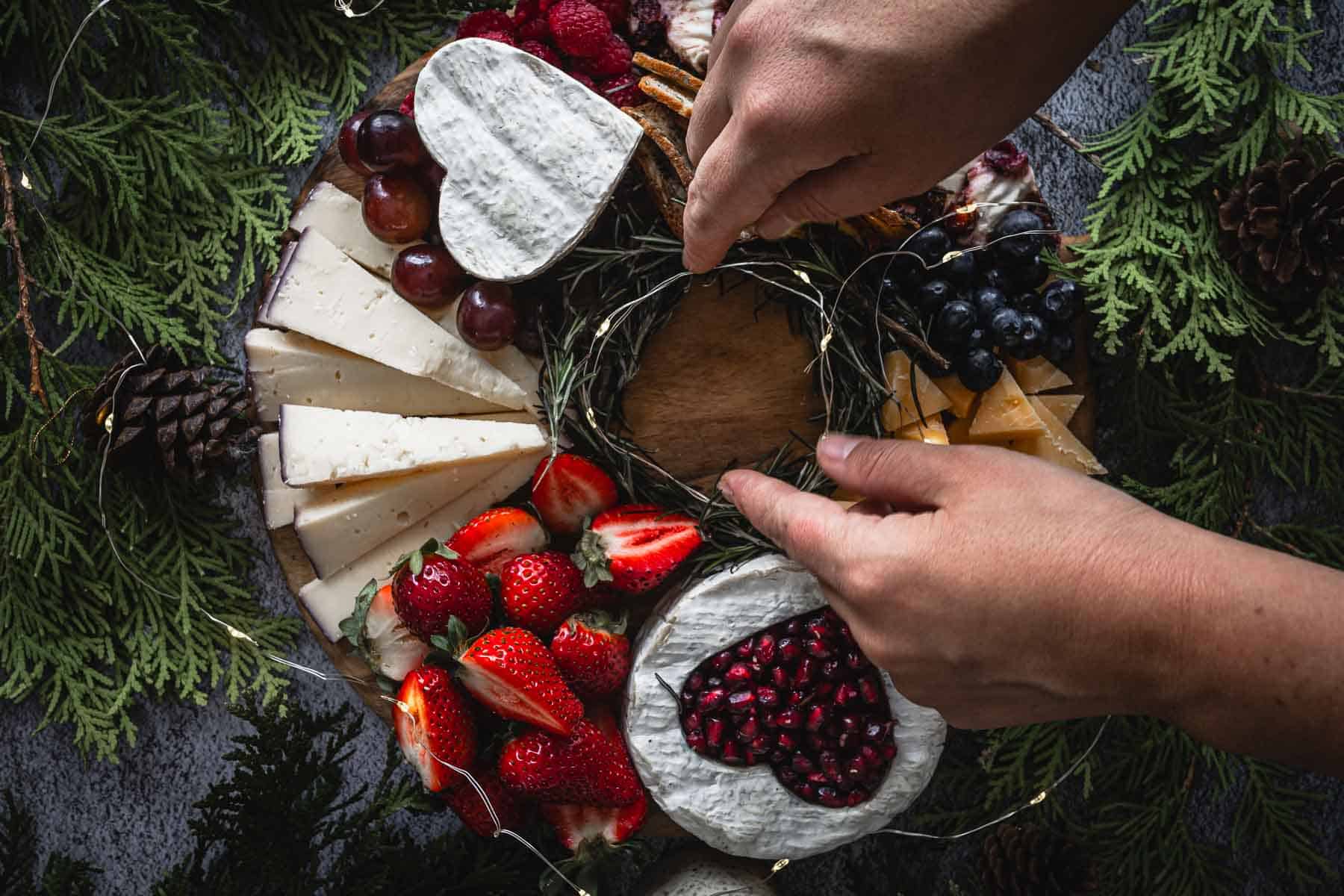 A person arranges a cheese platter adorned with various cheeses, strawberries, grapes, and blueberries. Two hands adjust decorations on the platter, which is surrounded by greenery and pinecones.