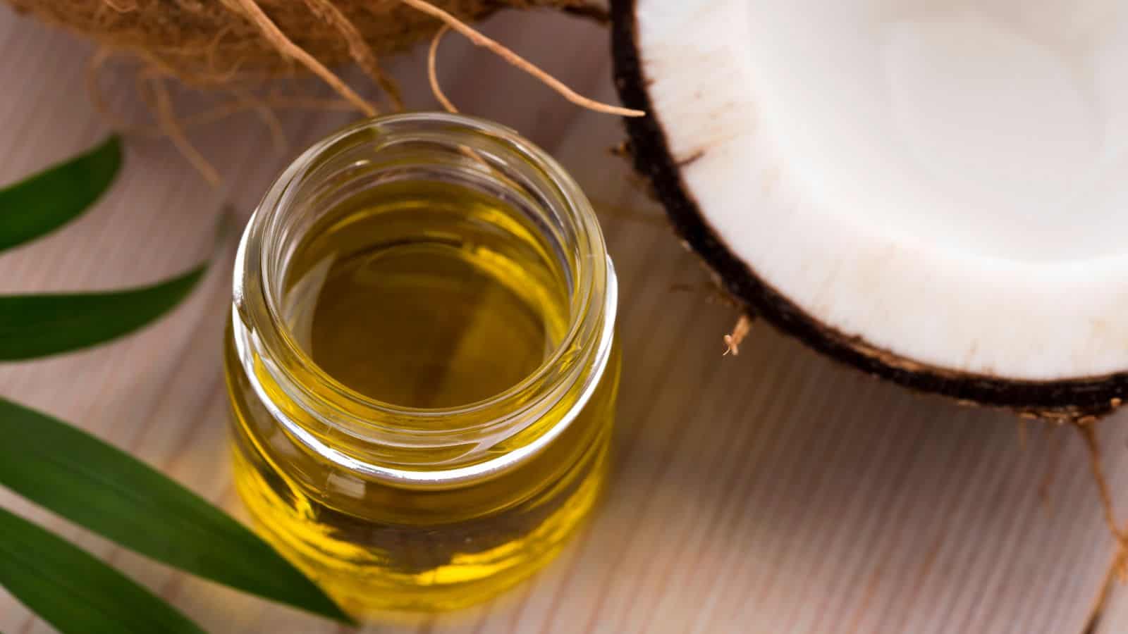 A jar of coconut oil is placed on a wooden surface next to half a coconut and green leaves. The oil is golden yellow, contrasting with the white flesh of the coconut. This image embodies a natural and organic theme, highlighting healthy oils in their purest form.