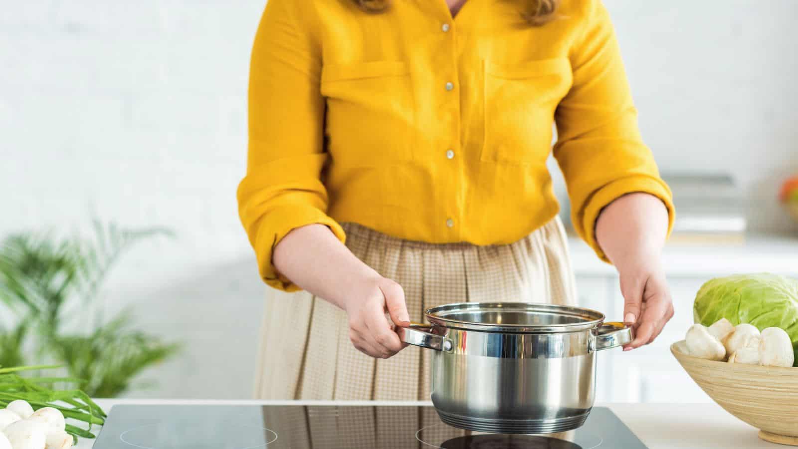 A person wearing a yellow shirt and beige skirt is holding a stainless steel pot over an induction cooktop in a kitchen. There is a bowl of vegetables, including a cabbage, on the counter nearby.