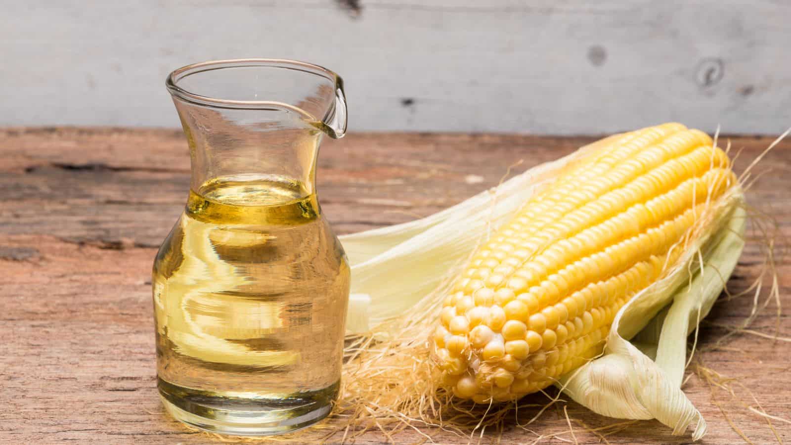 A glass pitcher filled with corn oil sits beside a fresh ear of corn, its husk partially peeled back, on a rustic wooden surface—highlighting the contrast between toxic and healthy oils.