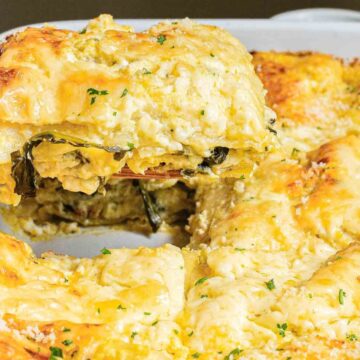 A close-up of a slice of creamy vegetable lasagna being lifted from a baking dish with a wooden spatula. The lasagna features layers of pasta, vegetables, and melted cheese, topped with parsley. The surrounding pieces show a golden-brown crust.