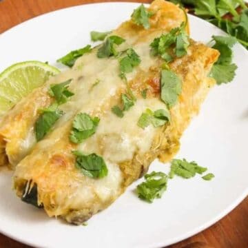A white plate with two enchiladas topped with melted cheese and cilantro, served alongside a slice of lime. The plate is placed on a wooden surface, with a bunch of cilantro nearby.