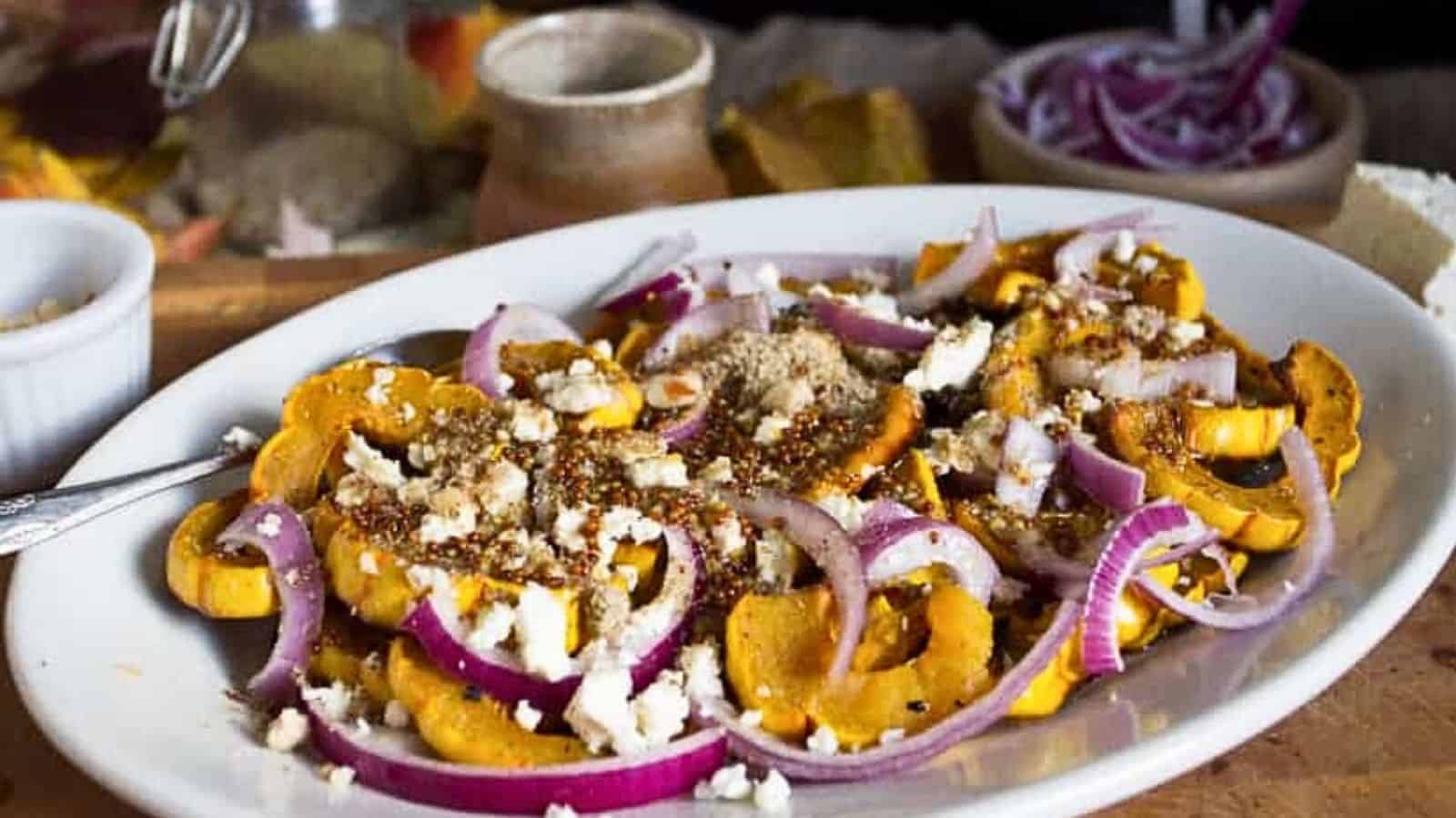 A white plate with roasted squash slices topped with red onion, crumbled cheese, and a sprinkle of nuts—this dish is bound to steal the spotlight. In the background, there are bowls and additional ingredients on a wooden surface. Perfect for insanely easy sides that impress.