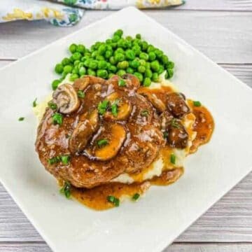 A square white plate holds mashed potatoes topped with a rich, brown mushroom gravy and slices, garnished with green herbs. A serving of green peas is on the side. Reminiscent of classic '60s recipes loved by Baby Boomers, a floral napkin peeks out beside the fork and knife on the left.